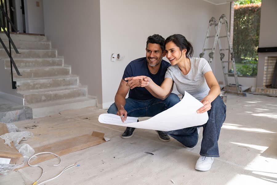 A smiling couple kneels on the floor of a house under home renovation, looking at blueprints together. The room features a ladder, stairs, exposed wiring, and unfinished surfaces.
