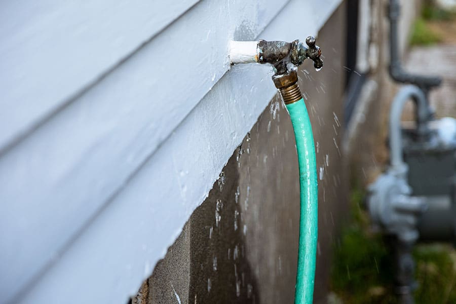 A green garden hose is attached to a leaky outdoor faucet on the side of a house, with water spraying out and running down the wall—an example of exterior problems that can lead to costly foundation damage if not addressed as part of regular home maintenance.