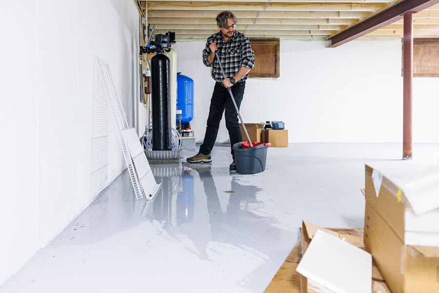 A man in a plaid shirt mops water from the floor of an unfinished basement, dealing with water problems. Nearby are boxes, shelving, and exposed ceiling beams.