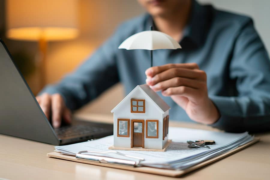 A person holds a small white umbrella over a model house on a desk, symbolizing homeowners insurance. The house, representative of Michigan homeowners seeking protection from risks like basement water damage, sits on a stack of papers beside a laptop and keys.